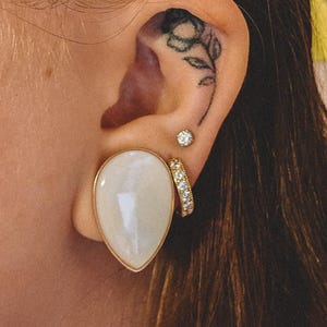 May include: A teardrop-shaped white stone ear plug with a gold-toned rim, accompanied by a small circular stud earring featuring a clear stone and a gold-toned hoop earring set with multiple small clear stones, worn in a pierced earlobe.
