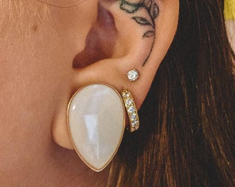 May include: A teardrop-shaped white stone ear plug with a gold-toned rim, accompanied by a small circular stud earring featuring a clear stone and a gold-toned hoop earring set with multiple small clear stones, worn in a pierced earlobe.