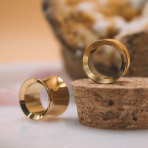 Puede incluir: Dos túneles para oreja de tono dorado pulido con una superficie lisa y reflectante y un diseño acampanado. Un túnel está colocado sobre una superficie plana, mientras que el otro descansa sobre un material de corcho marrón, cilíndrico y texturizado.