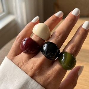 Peut inclure: Un ensemble de quatre bagues bombées, lisses et épaisses aux couleurs unies. Ces bagues présentent une finition brillante et sont portées sur quatre doigts, affichant une palette de couleurs crème, bordeaux foncé, noir et vert olive.