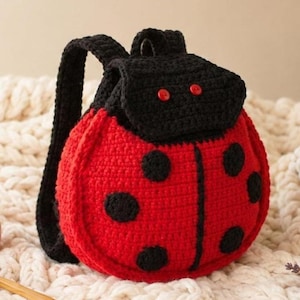 Puede incluir: Una mochila de ganchillo con forma de mariquita, cuerpo rojo brillante y puntos negros. La bolsa cuenta con una solapa negra con dos pequeños botones rojos, correas negras para los hombros y una franja vertical negra central.