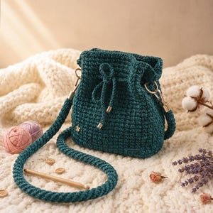 Può includere: Una borsa a secchiello all'uncinetto color verde ottanio scuro con chiusura a coulisse, dotata di una lunga tracolla abbinata, dettagli in metallo dorato e un piccolo ciondolo a forma di ape dorata sul davanti con un motivo a rilievo.