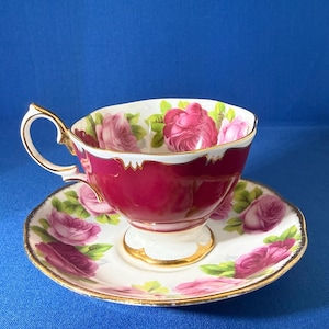 Puede incluir: Un juego de taza de té y platillo de porcelana con un exterior magenta intenso y un interior blanco. Ambas piezas están decoradas con motivos de rosas rosadas, hojas verdes y bordes dorados en los bordes festoneados y el asa.