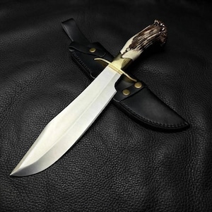 Peut inclure: Un couteau à lame fixe avec une lame en acier argenté poli, une garde en laiton et un manche en bois de cerf brun texturé. Le couteau est placé en diagonale sur un étui en cuir noir doté de deux rivets en laiton et d'un bouton-pression.