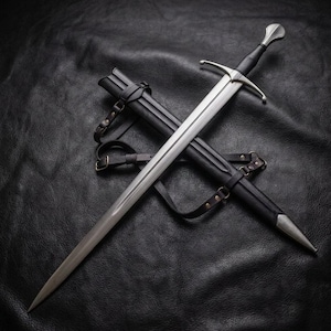 May include: A silver-toned steel sword with a double-edged blade, a cross-shaped guard, and a black leather-wrapped grip. The sword is positioned diagonally across a black leather scabbard that features matching black leather straps and metal buckles.