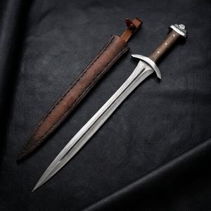 Könnte beinhalten: Ein zweischneidiger Stahldolch mit einer zentralen Hohlkehle, ausgestattet mit einem dunklen Holzgriff mit silberfarbenen Metallakzenten und einem Knauf. Der Dolch wird von einer braunen Lederscheide mit Ziernähten und einem Riemenverschluss begleitet.