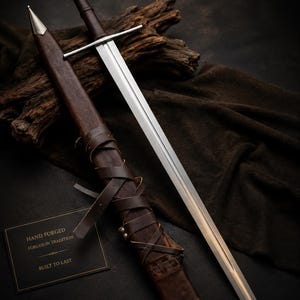 Pode incluir: Uma espada de aço com guarda reta e punho revestido em couro castanho escuro, acompanhada por uma bainha de couro castanho com atacadores. Um cartão preto apresenta o texto: HAND FORGED, FORGED IN TRADITION, BUILT TO LAST.