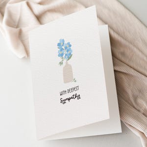 May include: A white folded greeting card featuring a minimalist illustration of light blue flowers in a beige vase. Below the vase, the text reads "WITH DEEPEST SYMPATHY" in a simple black sans-serif and script font combination on the front cover.