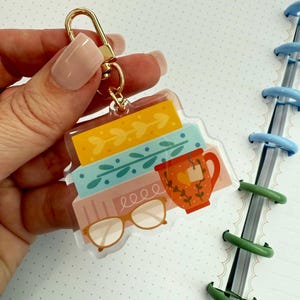 May include: A clear acrylic keyring with a design of books, a teacup, and spectacles. The books are yellow, blue, and pink. The teacup is orange with a heart. The keyring has a gold-coloured clasp.
