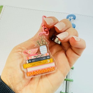 May include: A clear acrylic keyring featuring a stack of books, a teacup, and a vase with plants. The books have colourful spines with decorative patterns. The keyring is attached to a pink ring.