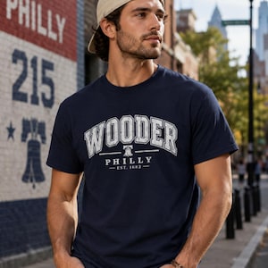 Könnte beinhalten: Ein marineblaues Kurzarm-T-Shirt mit weißem, abgenutztem Text, der WOODER PHILLY EST. 1682 lautet, sowie einem kleinen Liberty Bell-Symbol zwischen den Wörtern. Das T-Shirt hat einen Rundhalsausschnitt und eine normale Passform.