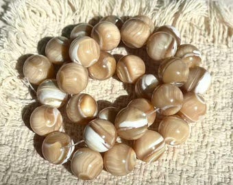 May include: A collection of round, smooth beads featuring a swirling pattern of tan, beige, and white colors. The beads have a glossy, polished finish and are arranged in a cluster, showing variations in the marbled light brown and cream tones.