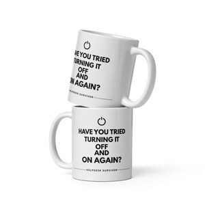 Könnte beinhalten: Zwei vertikal gestapelte weiße Keramiktassen. Jede Tasse ist mit einem schwarzen Power-Symbol, dem Text "HAVE YOU TRIED TURNING IT OFF AND ON AGAIN?" und dem Schriftzug "HELPDESK SURVIVOR" über einer horizontalen Linie bedruckt.