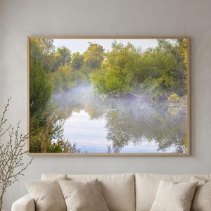 Può includere: Una grande stampa artistica rettangolare da parete che raffigura un paesaggio sereno di alberi verdi lussureggianti riflessi in un fiume calmo e nebbioso. L'opera è incorniciata in legno chiaro sopra un divano beige con cuscini abbinati.