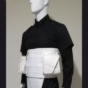 Op de afbeelding: Een zwarte set met een shirt met lange mouwen en een broek, voorzien van een witte heupriem met twee zijzakken en een wit kruisstuk. Het ensemble bestaat uit een zwart shirt met een hoge kraag en een bijpassende zwarte broek.