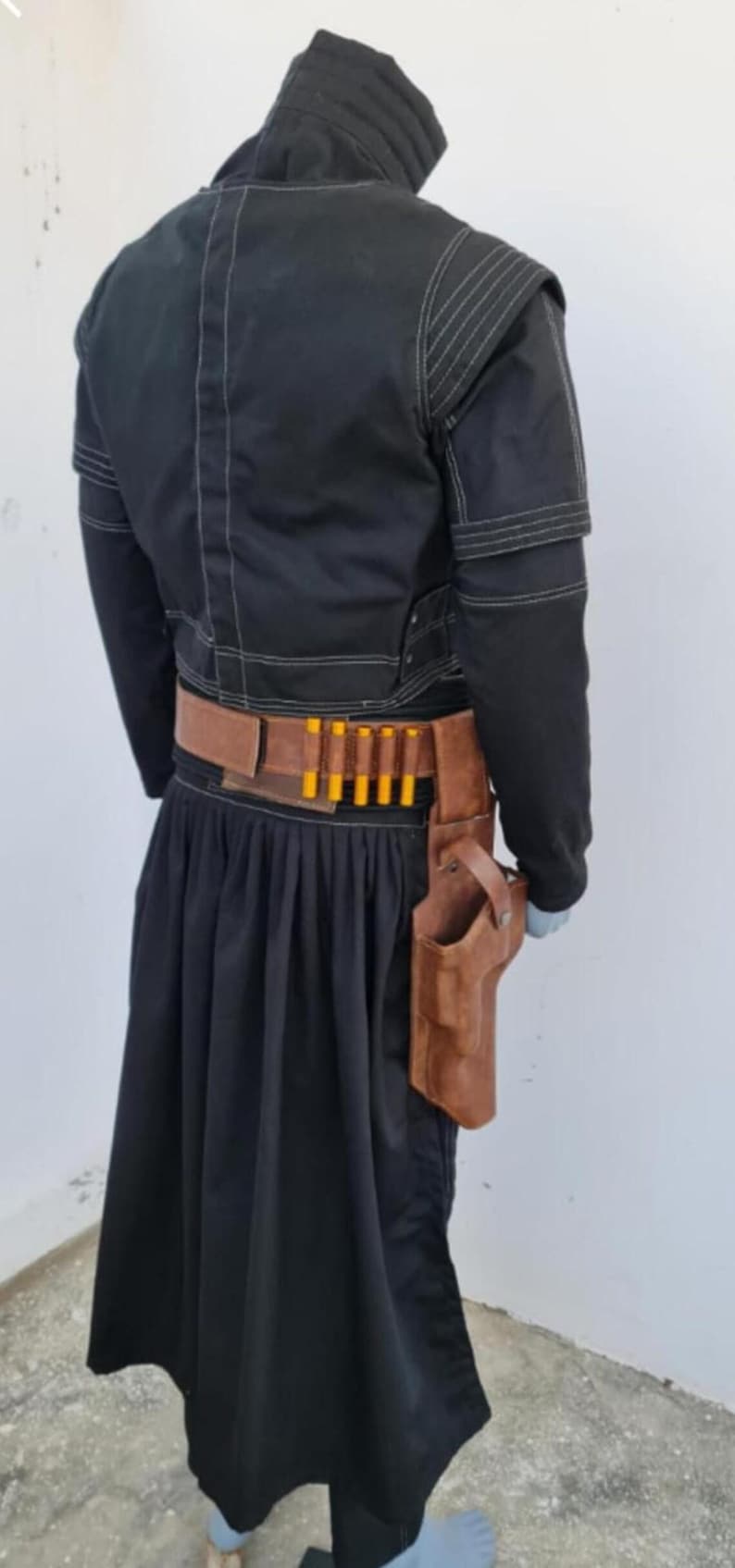 May include: A black jacket with white stitching and a high collar, paired with a black pleated skirt. A brown leather belt features a row of five yellow-tipped cartridges and a matching brown leather holster attached to the side of the belt.