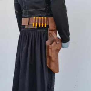 May include: A black jacket with white stitching, a black pleated skirt, and a brown leather belt featuring a holster and five yellow-tipped cartridges. The ensemble is displayed on a mannequin, showcasing the layered construction and utility-style design.