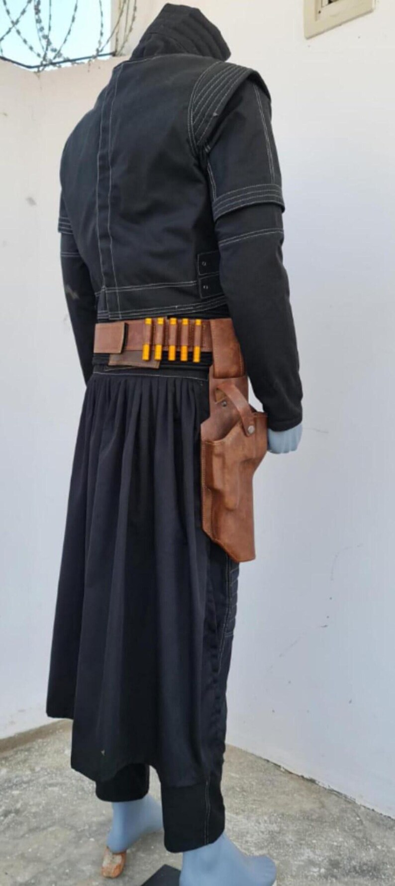 May include: A black jacket with white stitching, a black pleated skirt, and a brown leather belt featuring a holster and five yellow-tipped cartridges. The ensemble is displayed on a mannequin, showcasing the layered construction and utility-style design.