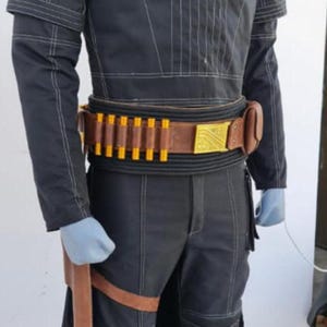 May include: A black long-sleeved jacket and matching trousers with white contrast stitching, a high collar, and ribbed shoulder and knee panels. A brown leather belt features a gold rectangular buckle and yellow cylindrical inserts.