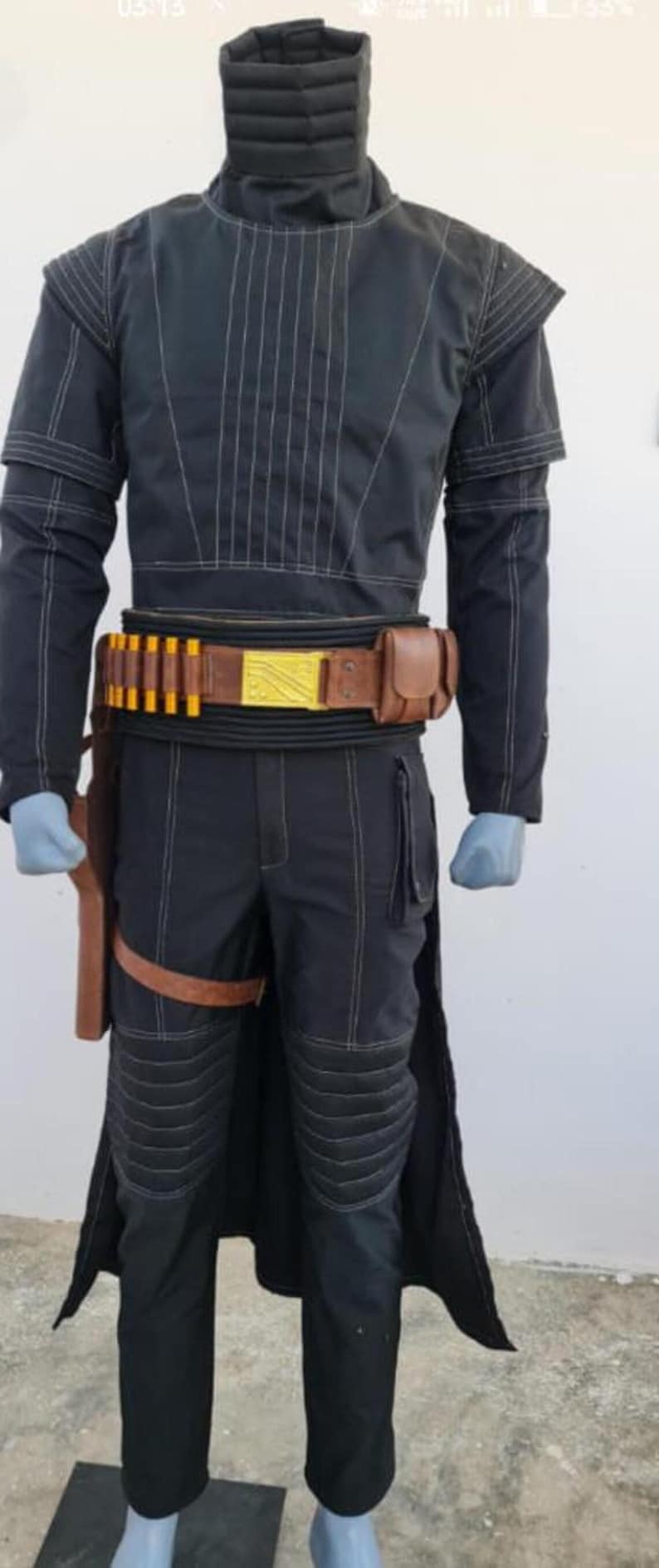 May include: A black long-sleeved shirt and matching pants with white topstitching, featuring a high neck collar, padded knee sections, and a brown leather utility belt with yellow cylindrical accents, a gold rectangular buckle, and a side holster.