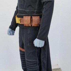 May include: A black long-sleeved tunic and trousers set featuring white contrast stitching, a high collar, and ribbed detailing on the shoulders and knees. A brown leather utility belt with two pouches and a gold-colored buckle is worn at the waist.