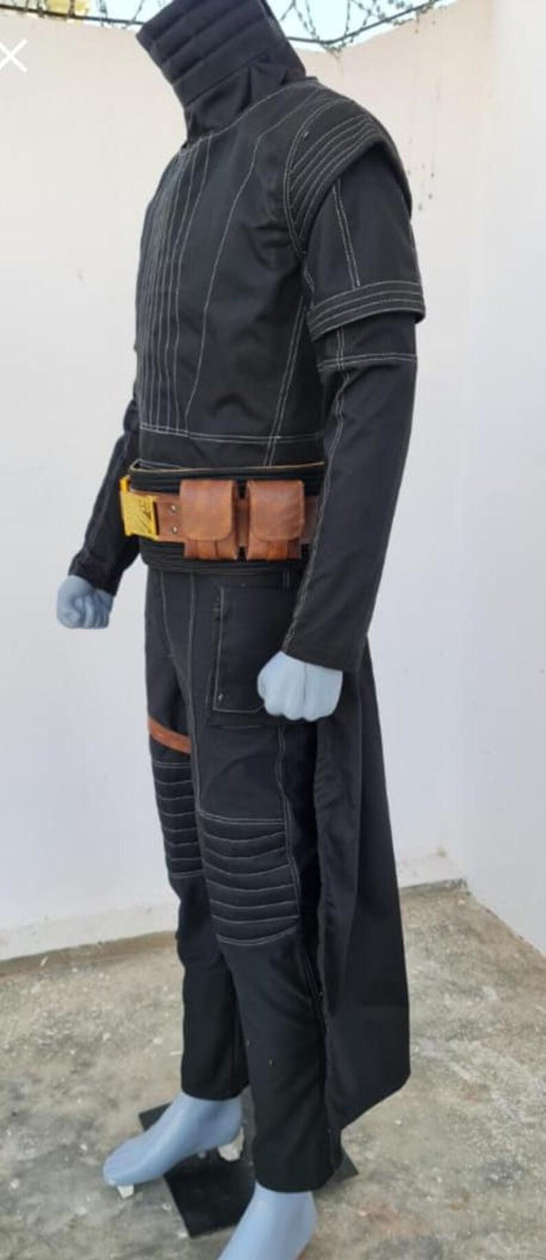 May include: A black long-sleeved tunic and trousers set featuring white contrast stitching, a high collar, and ribbed detailing on the shoulders and knees. A brown leather utility belt with two pouches and a gold-colored buckle is worn at the waist.