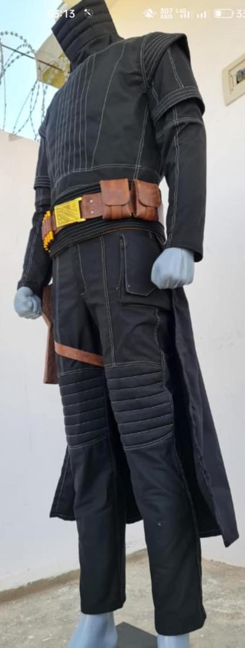May include: A black long-sleeved tunic and matching trousers featuring white contrast stitching, a high collar, and quilted knee panels. The outfit includes a brown utility belt with pouches, a gold-colored buckle, and a brown leg holster strap.