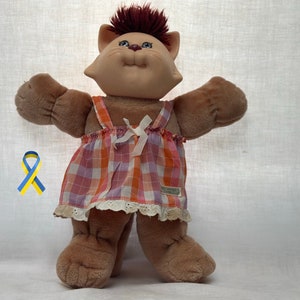 May include: A brown plush cat doll wearing a pink and orange checked dress with a white lace trim. The doll has a brown nose, blue eyes, and a red mohawk.