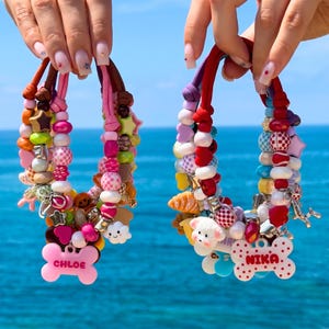May include: Two beaded pet collars featuring colorful plastic beads in various shapes, including stars, hearts, and smiley faces. One collar has a pink bone-shaped tag with the name CHLOE, and the other has a white bone-shaped tag with the name NIKA.