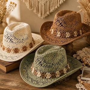 Peut inclure: Trois chapeaux de cowboy en crochet de couleur crème, marron et vert sauge. Chaque chapeau présente un motif à mailles ouvertes et une bande décorative composée de petites fleurs en crochet multicolores autour de la base de la calotte.