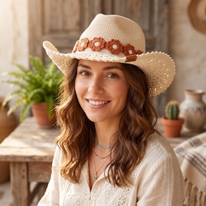 May include: A cream-colored woven hat featuring a wide brim and a decorative band of crocheted rust-colored flowers. The hat has a structured crown and a small brown rectangular tag on the side. The material appears to be a textured straw weave.