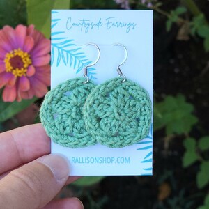 Hand holds out a card with a set of crochet green flower earrings on it.