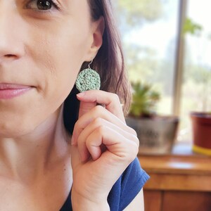 A green flower earring is modeled on a woman holding it out for size reference.