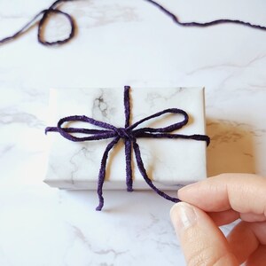 Gift wrapped earrings are in a jewelry gift box with a white marble design. A purple velvet ribbon is tied around the box in a bow.