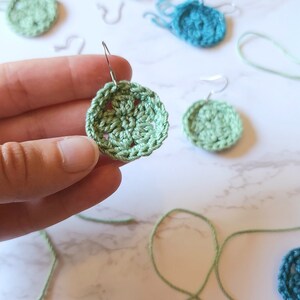 A hand holds out a green crochet flower earring with several unfinished earrings in the background along with a crochet hook and yarn.