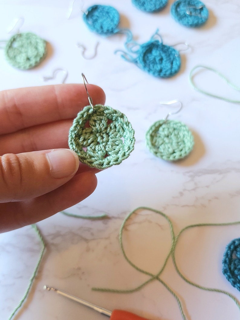 A hand holds out a green crochet flower earring with several unfinished earrings in the background along with a crochet hook and yarn.