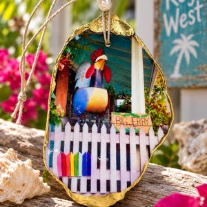 May include: An oyster shell ornament with a gold-painted edge, featuring a printed image of a colourful rooster statue behind a white picket fence with a small rainbow section and a sign that reads BIG JERRY, suspended by a twine cord with beads.