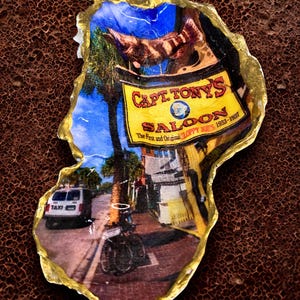 Könnte beinhalten: Eine Austernschale mit Decoupage-Motiv, das eine lebhafte Straßenszene von Capt. Tony's Saloon mit einem gelben Schild, einem weißen Taxi und Palmen zeigt. Die Schale hat einen goldfarbenen Rand und eine glänzende Oberfläche mit dem Text Capt. Tony's Saloon.