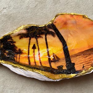 May include: An oyster shell painted with a sunset scene featuring black silhouettes of palm trees and a sailboat against a vibrant orange and yellow sky. The edges of the shell are trimmed with a metallic gold paint, highlighting the natural texture.