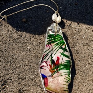 May include: A decorative hanging ornament made from a translucent, irregular shell-shaped piece featuring a printed image of roosters among green palm fronds. The piece is suspended by a thin, light-coloured string adorned with two small white beads.