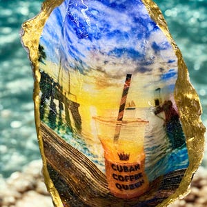 Puede incluir: Una concha de ostra con bordes pintados en oro que presenta una imagen impresa de un atardecer sobre un muelle y agua. Un vaso de plástico con una pajita a rayas está en primer plano, con un icono de corona y el texto CUBAN COFFEE QUEEN.