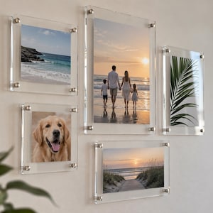 May include: A set of five clear acrylic floating photo frames mounted on a wall. The frames display various images including a beach landscape, a family walking on the shore at sunset, a golden retriever, a palm leaf, and a coastal path at dusk.