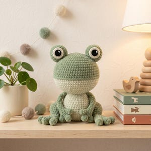 Puede incluir: Un peluche de rana hecho a ganchillo con cabeza y parte superior del cuerpo de color verde salvia y una barriga de color crema más claro. La rana tiene ojos grandes y redondos con pupilas negras y reflejos blancos, y cuatro patas extendidas.
