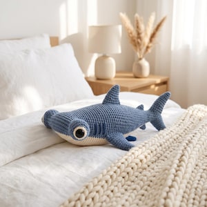 May include: A blue and cream-coloured crochet hammerhead shark plush toy. The shark features a wide, flat head with large, round black eyes, a dorsal fin, pectoral fins, and a tail fin. The body is constructed with visible yarn stitches.