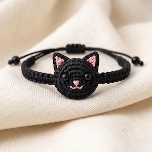 Puede incluir: Una pulsera de macramé negra con una cara de gato tejida a ganchillo en el centro. El gato tiene detalles en hilo negro, orejas interiores bordadas en rosa, nariz y boca rosas, y dos pequeños ojos de cuentas negras como adorno.