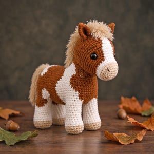 May include: A crocheted horse toy featuring a brown and white spotted coat, a cream-colored mane and tail, and dark plastic eyes. The horse stands on four legs with light tan hooves, showcasing detailed yarn stitching throughout its body.