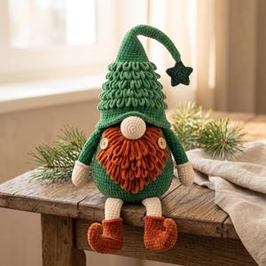 Puede incluir: Un muñeco gnomo de ganchillo con un sombrero verde texturizado con una estrella verde oscuro colgante, una nariz color crema, una barba naranja espesa y un cuerpo verde. El gnomo tiene brazos y piernas color crema con zapatos naranjas de punta curva.
