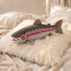 Peut inclure: Un poisson en peluche crocheté avec un dos gris foncé, un ventre gris clair et une bande rose horizontale distincte sur le côté. Le poisson présente une surface en laine texturée, une petite bouche ouverte et un œil marron visible.