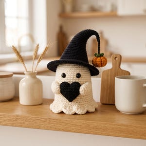 Peut inclure: Une figurine de fantôme crochetée en fil blanc cassé, portant un grand chapeau pointu noir avec une petite citrouille orange suspendue au bout. Le fantôme a deux yeux en perles noires et tient un cœur crocheté noir contre son corps.