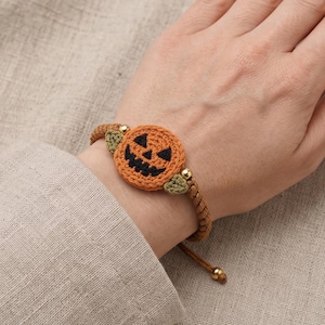 May include: A bracelet featuring a crocheted orange pumpkin charm with a black stitched jack-o'-lantern face, accented by two small green crocheted leaves, gold-toned metal beads, and a brown braided cord band with adjustable sliding knots.